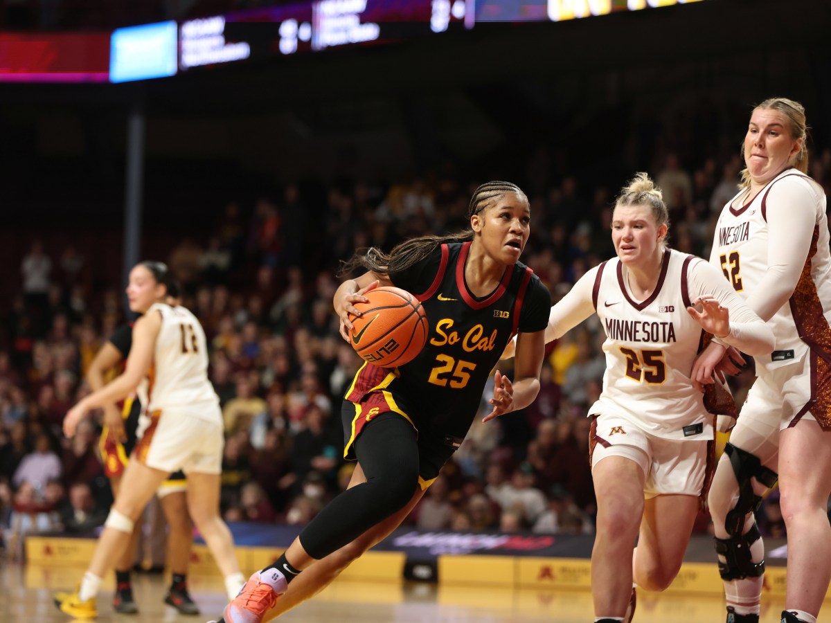 USC Trojan forward Kara Dunn dribbles down the court past a pursuing Minnesota Gopher forward Grace Grocholski, who is trying to get through her teammate Sophie Hart. Another Gopher stands away near midcourt. In the background and out of focus is a packed arena crowd.