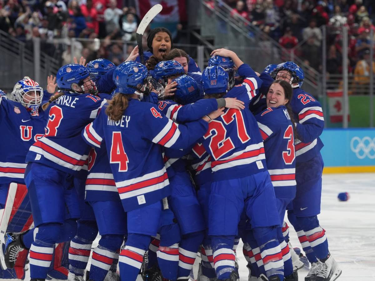 Team USA celebrates their gold medal win with smiles and a big, tight group hug at center ice. They are all wearing blue home uniforms.